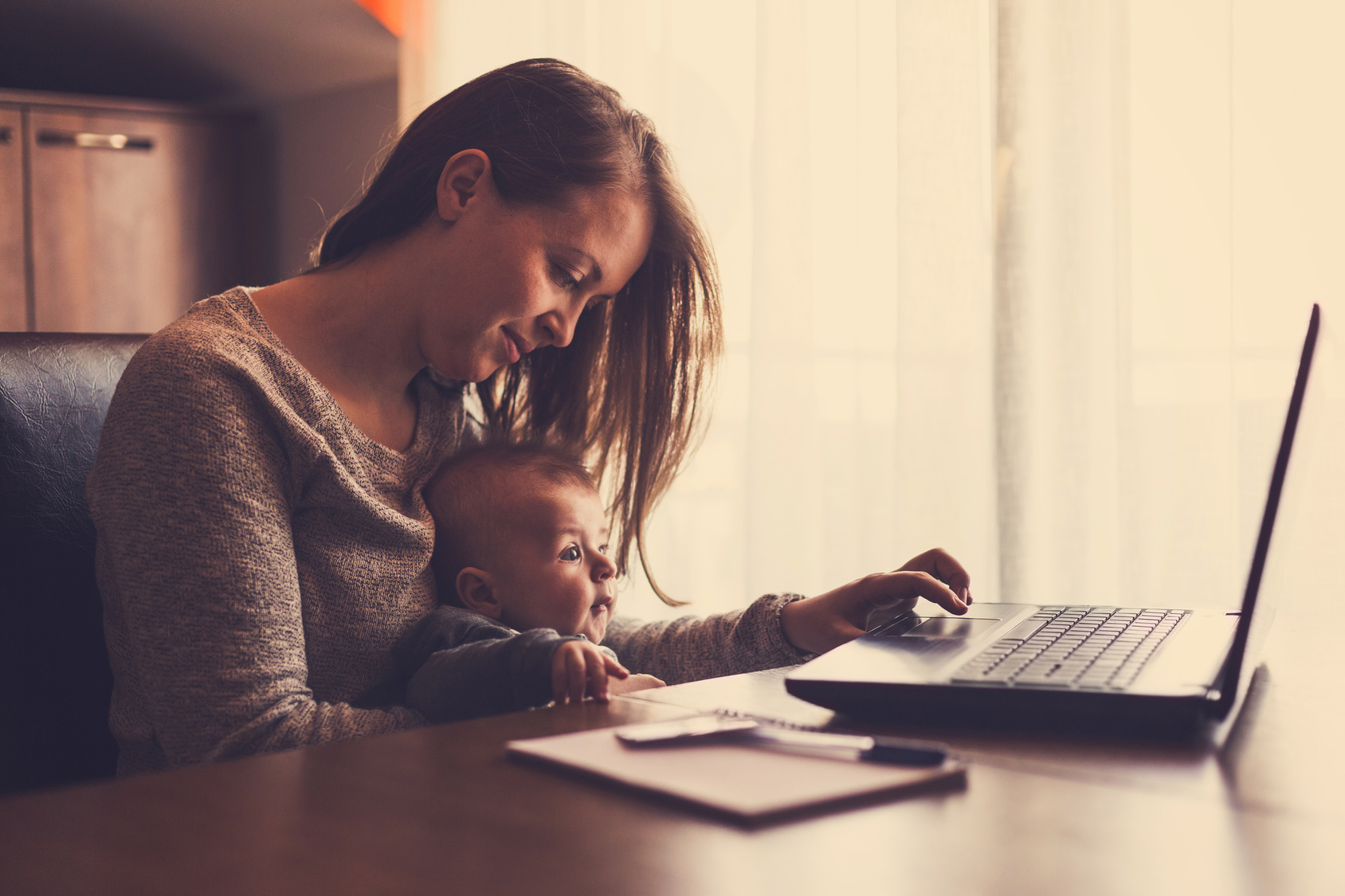Young single mother searching for a bloomfield hills divorce lawyer on a laptop computer at home. She is holding her baby boy in her arms and sitting at the kitchens table.